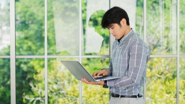 Asyalı işadamı gülümsüyor ve ofisin penceresinin yanındaki dizüstü bilgisayarda çalışıyor. Ofis lobisinde bilgisayarla buluşan yakışıklı bir adamın portresi.