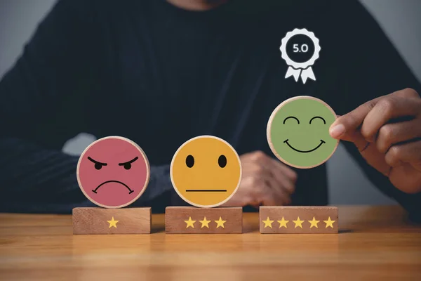 Hand choosing a smiley face on a wooden block circle to rate the best business services and customer experience. Satisfaction survey and 5-star rating concept.