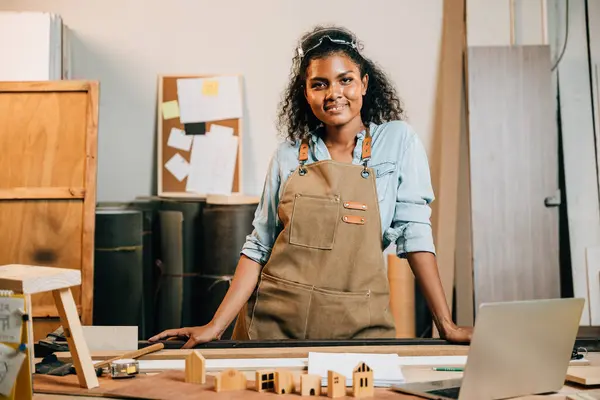 Portrait of american black woman curly hair carpenter smile working in carpentry DIY on workspace table at workshop against wood or woodshop, industry, Happy Carpenters Day