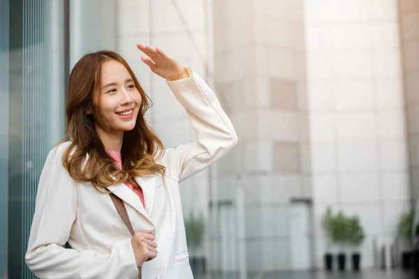 Happy woman wearing white suit jacket with brown bag at sunlight outdoors. Portrait of confidence young businesswoman standing outside office building in city raise your hand to shade sun