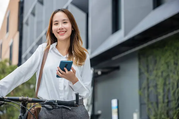 在城市里用智能手机在户外换乘自行车 在办公室里骑自行车 站在城市街道上骑自行车的亚洲女商人拿着手机 — 图库照片