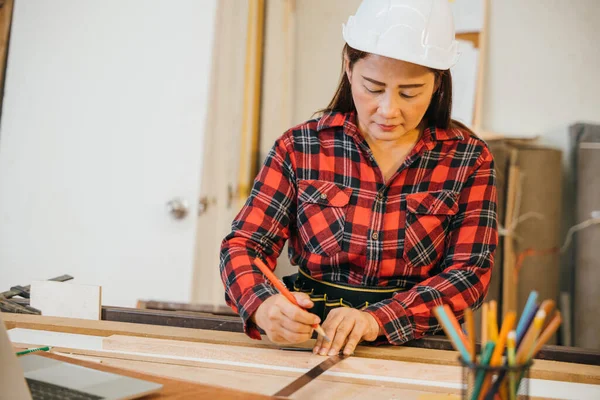 Asian senior woman carpenter taking measurement of a wooden plank and make in workshop, female holding ruler and use pencil drawing sign on plank at woodshop, National Carpenters Day