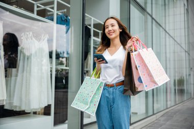 Asian beautiful woman holding shopping bag and smartphone on her hands near the mall shop window, Smiling shopper young girl standing using mobile phone after shopping on city street