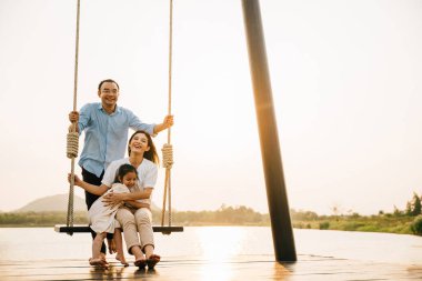 Mutlu bir aile parkta güneşli bir günün tadını çıkarıyor, baba oturuyor ve salıncakta salıncak sallarken anne ve kız birlikte oynuyorlar, Mutlu Aile Günü