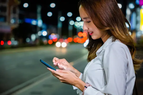 Portrait of a Young Asian woman on the streets of downtown. She using mobile app device on smartphone in downtown city street, with illuminated busy city traffic scene during rush hour.