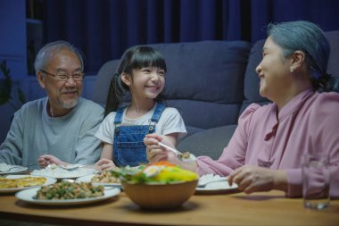 Mutlu Asyalı aile büyükbabası ve torunu akşam yemeği yerken ve evde eğlenirken, kıdemli ebeveyn ve çocuk oturma odasında birlikte akşam yemeği yerken.