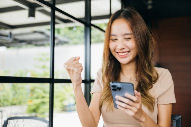 Asyalı genç bir kadın iş başarısını cep telefonuyla kutluyor, güzel bir kadın dışarıda akıllı telefon kazanan bir jestle el ele tutuşuyor.