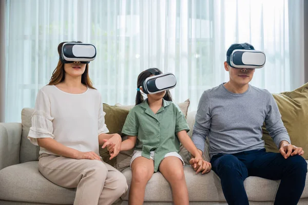 Happy family have mother father and daughter watching movie or playing video game, game entertainment innovation technology, Asian family wear vr glasses headsets excited sitting sofa in living room