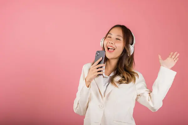Woman excited smiling listening to music radio in bluetooth headphones and singing song on smartphone studio shot isolated pink background, happy Asian young female Karaoke online app in mobile phone