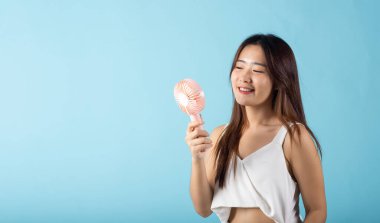 Asian beauty young woman holding pink portable electric mini fan near her face studio shot isolated on blue background, Female hand hold small plastic fan handheld she enjoying cool wind blowing