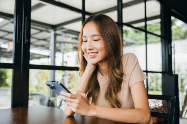 Asyalı genç bir kadın, sabah kafede online alışveriş yapmak için akıllı telefon kullanıyor. Kadın, cep telefonundan para transferi yapıyor.