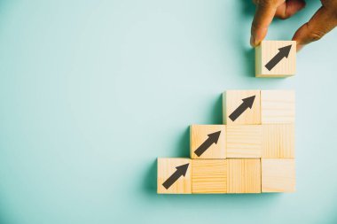 Erkek eller tahta blokları beyaz bir masaya basamaklar gibi diziyor. Yukarı bakan bir okla iş büyümesi kavramı, gelecekteki gelişmeyi ve hedeflere ulaşmayı sembolize ediyor. Boşluğu kopyala.