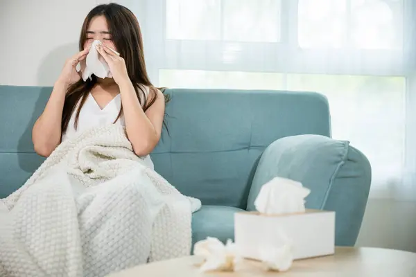 Sick female sitting under blanket on sofa and sneeze with tissue paper in living room, fever caught cold, Asian young woman she sick blowing nose sneezing in tissue at home, medicine healthcare