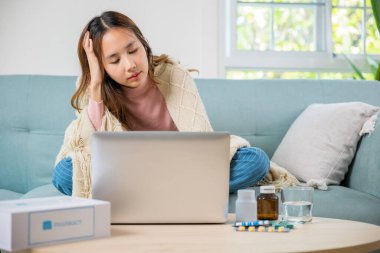 Sick young business woman sit on sofa suffering from cold covered blanket still work with laptop computer, influenza symptoms, Unwell female working online feeling sick unhealthy, medicine on desk