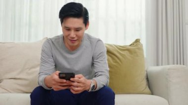 Asyalı, orta yaşlı bir adam koltukta oturmuş akıllı cep telefonu kullanıyor, mutlu, olgun erkekler sosyal medyada gülümsüyor oturma odasında akıllı telefondan sohbet ediyor, elinde bir cihaz tutuyor.