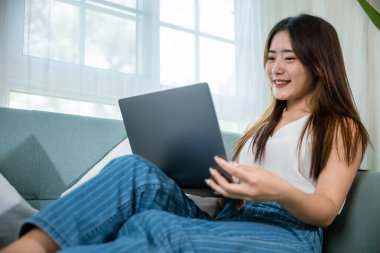 Oturma odasında dizüstü bilgisayar kullanarak koltukta oturup gülümseyen Asyalı genç bir kadın bilgisayar projesinde çalışan mutlu bir kadın dizüstü bilgisayarda e-posta yazıyor.