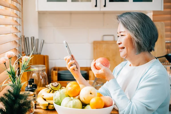 In the kitchen, an old Asian woman, a grandmother, enjoys her breakfast. She smiles while using her smartphone and eating a red apple. A portrayal of modern technology and happiness.