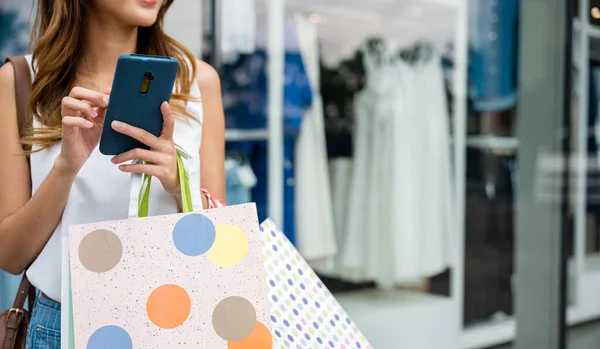 ショッピングバッグと携帯電話を持つ女性は 最新のトレンドのために買い物しながら 技術の利便性を楽しみ 店の窓の前に立っています ソーシャルメディアを使用して現代の消費者を共有 — ストック写真