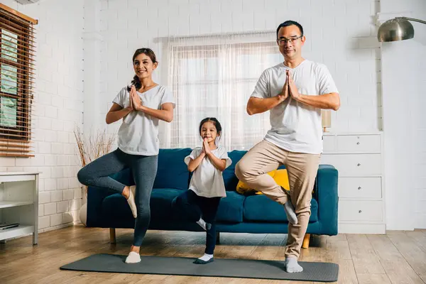 A family of three Asian parents and their daughter focus on yoga and fitness training at home fostering health happiness and togetherness.