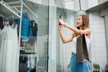Moda mağazasının önünde elinde kağıt torbalarla akıllı telefonunu karıştıran güzel bir genç kadın. Çevrimiçi alışveriş ve sosyal medyanın dijital dünyasına bağlı..