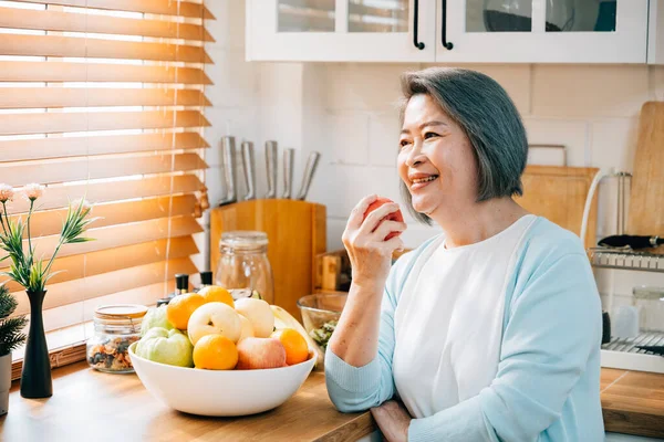 In kitchen at home, smiling old Asian woman grandmother enjoys cooking and preparing healthy vegan food. Her joy is evident as she chooses a fresh apple to eat. portrait of diet concept and happiness.