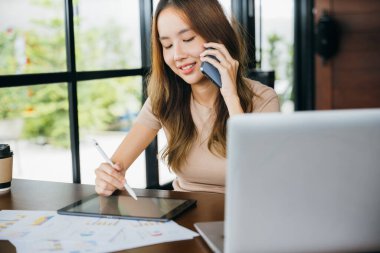 Kafe 'de oturan Asyalı iş kadını cep telefonuyla konuşuyor ve bir şeyler soruyor, gülümseyen bir kadın bilgisayar çalışıyor ve evdeki dijital tablete not alıyor.