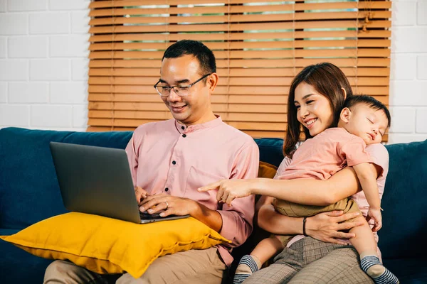 In their home office a husband works on his laptop while his wife carries their sleeping daughter. This portrait captures the essence of balancing work family and responsibility.