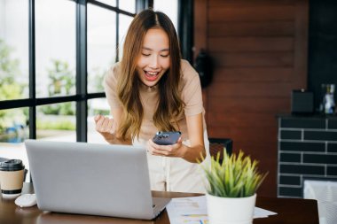 Heyecanlı Asyalı genç bir kadın cep telefonuyla kutlama yapıyor. Mutlu bir başarı, akıllı telefondan iyi haberler alıyor. Cefa 'da bilgisayar ve sosyal medya ağında çalışırken.