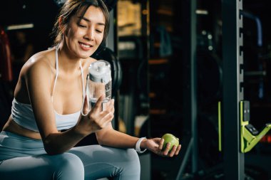 Spor salonunda elinde yeşil elma ve su şişesi tutan gülümseyen genç bir kadın, sağlıklı gıda seçenekleri ve fitness hedeflerine ulaşmak için susuzluğun önemini vurguluyor. sağlıklı yaşam tarzı vücut geliştirme