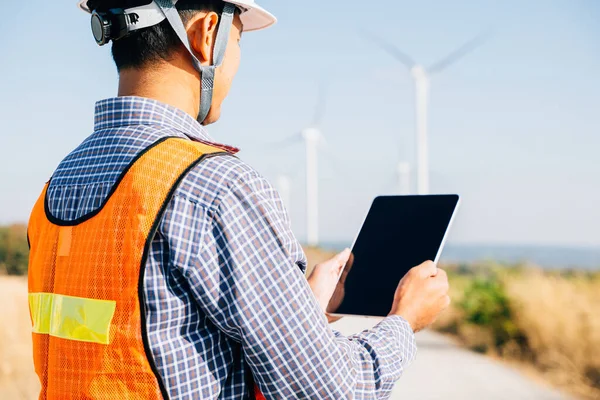 An engineer tablet in hand works at windmill farm. Generating clean energy the technicians expertise guarantees turbine efficiency. Global innovation in electricity control a confident stance.