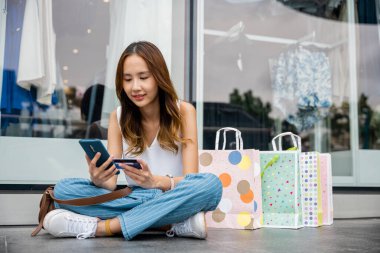 Perakende terapiden hoşlanan bin yıllık bir kadın elinde alışveriş torbaları, kredi kartı ve akıllı telefonuyla yerde oturuyor. Arka planda bir dükkan penceresi var. Alışveriş onun rahatlığıdır..