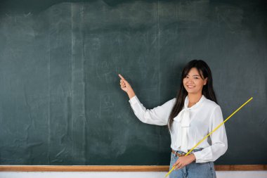Back to school concept. Happy beautiful young woman standing hold pointer to back board, Asian female teacher smiling with wooden stick pointing to blackboard at school in classroom, Education