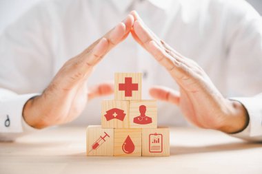 Sağlık simgeleriyle ahşap küp bloğu ve koruma işareti gösteren bir doktor eli, sağlık ve sigorta konseptini tasvir ediyor. Güvenli bir gelecek için tıbbi hizmetler.
