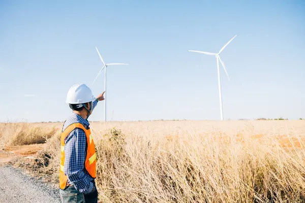 Man engineer in hardhat checks windmill farm. Generating clean electricity an Asian technician ensures turbine efficiency. Expertise in action innovating for global energy security.