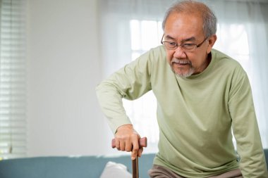 Diz ağrısı çeken yaşlı bir adam baston tutarken, yaşlı bir adam kanepeden tek başına kalkmak için yazı yazıyor bastonla eve yürümek için, emeklilik sağlık sigortası konsepti