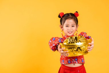 Çin Yeni Yılı. Mutlu Asyalı küçük kız gülümsemesi kırmızı Cheongsam 'ın elinde büyük altın külçeler, geleneksel elbiseli çocukların portresi altın külçeler, sarı arka planda kısa bir stüdyo.