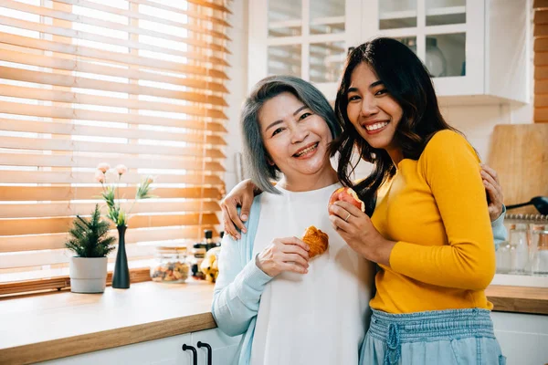 An elderly mother and her adult daughter, a cheerful young woman, hold an apple in the kitchen. Their smiles convey the joy of teaching, learning, and family togetherness.