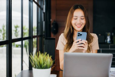 Kahve dükkanında akıllı telefondan gülümseyen mutlu bir kadın dizüstü bilgisayarla çalışan genç bir iş kadını Cefa 'da mesaj atmak için akıllı cep telefonu tutuyor.