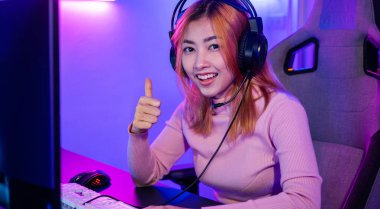 Oyun oynayan oyuncu oyun kulaklığı takıyor kameraya bakıyor. Başparmak işareti vererek başarıyı ifade ediyor. Gülümseyen kadın canlı yayında evde neon ışıkları altında video oyunu oynuyor.