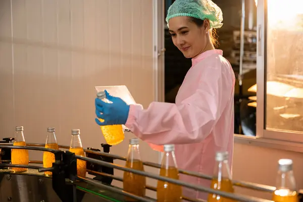 Woman technician in uniform checks bottles on conveyor using laptop. Quality control manager ensures drinks perfection. Science meets manufacturing as technology-driven control is maintained.