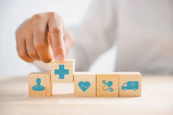 Hand gripping wooden block with healthcare and medical icons. Depicts safety, health, and family well-being, symbolizing pharmacy, heart care, and happiness. health care concept