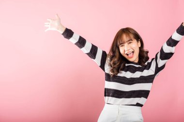 Asyalı bir kadın gülümsüyor, yumruklarını kaldırarak zafer jesti yapıyor, başarısını kutluyor. Pembe bir arka planda stüdyo çekimi, kazananın neşesini ve mutluluğunu yakalıyor..