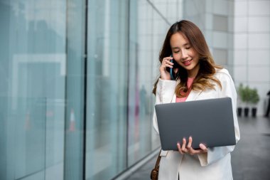 Portre güzel iş kadını gülümsüyor elinde bilgisayarla şehirdeki akıllı cep telefonuyla sorunları tartışıyor, Asyalı iş kadını dizüstü bilgisayarla çalışıyor ve ofisin yakınındaki cep telefonuyla konuşuyor.