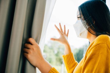 Koruyucu maske takan üzgün bir Asyalı kadın pencereden dışarı bakar COVID-19 yüzünden kendini yalnız ve sıkıntılı hisseder. Korku izolasyonunu ve pandemik anksiyeteyi yansıtıyor..