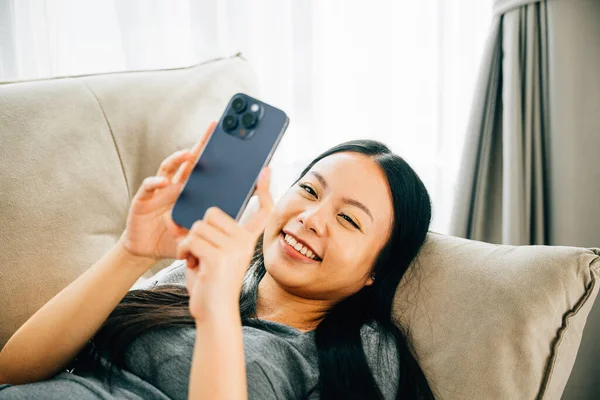 Young Asian woman relaxes on sofa texting on smartphone. Enjoying relaxation chatting and online shopping. Modern technology for communication and connection.