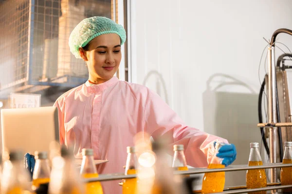 In a beverage factory a woman engineer conducts quality checks on bottles moving on a conveyor belt. Using a laptop she ensures stringent liquid standards.