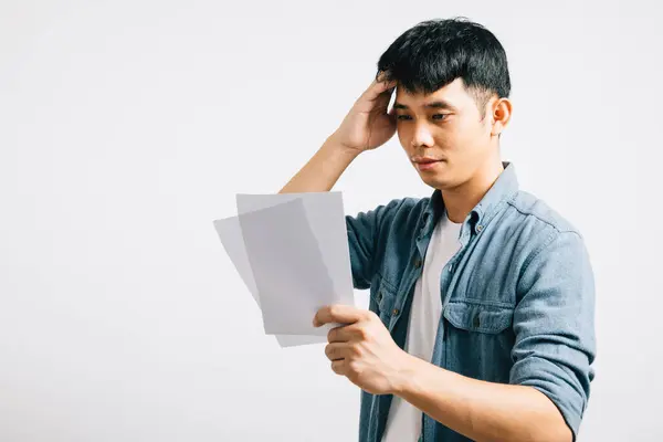 Worried over bills, an Asian man is portrayed in a studio shot, holding a document and appearing confused, emphasizing financial stress and trouble on a white background. over bill