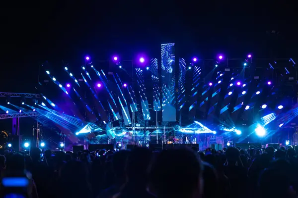 A lively concert festival at night with a cheering unrecognizable crowd gathered in front of the brightly lit stage. The lens flare adds to the excitement as people have blast during this music event.