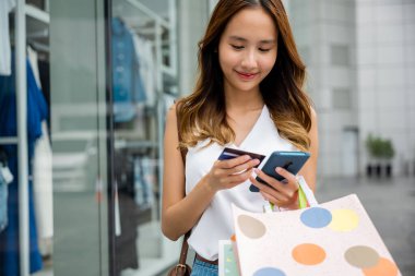 Alış veriş yapmak için akıllı telefonunu kullanan ve elinde alışveriş torbaları ve kredi kartı tutarken bağlı kalan tüketici ve genç kadın. Teknoloji ve alışveriş en iyisi.