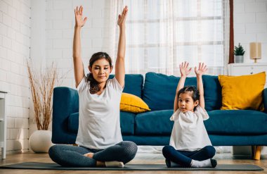 Güzel bir genç kadın ve büyüleyici kızı, evlerindeki yoga seanslarında gülümseyerek esnek ve dengeli bir ortam yaratıp uyumlu ve neşeli bir an yaratıyorlar..